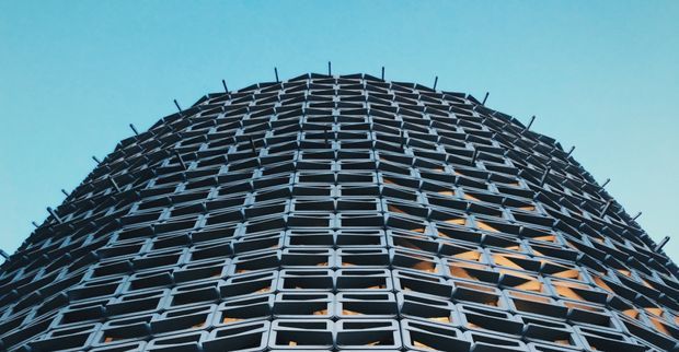 Upward-looking architectural photograph of a tall, rounded building facade covered in a dense repeating grid of projecting rectangular window or balcony frames. The dark metal-like facade contrasts strongly with a vivid blue sky, while warm interior light is visible through some openings. The image emphasizes the building's curved form and repetitive geometric pattern.