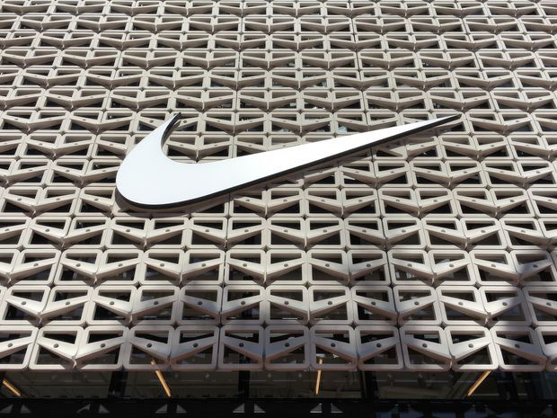 Close-up exterior view of a contemporary commercial building facade featuring a dense repeating geometric sun-screen or brise-soleil pattern in light grey, with a large white Nike swoosh mounted prominently across the surface. Dark glazing and structural elements are visible along the lower edge.