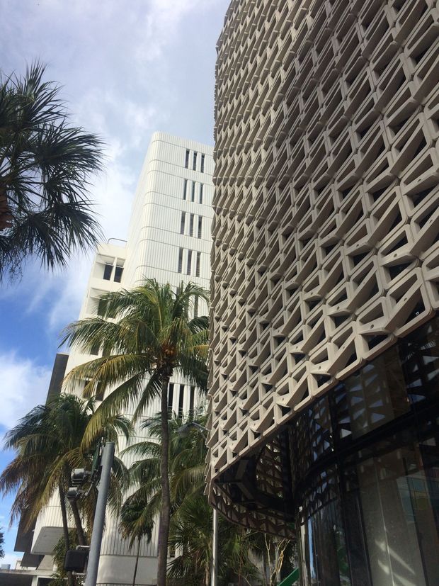 Upward-looking urban exterior view of a tall contemporary building with a prominent repeating geometric concrete or precast facade screen. Dark glazed frontage is visible at ground level, with a lighter modern tower behind it and palm trees lining the streetscape.