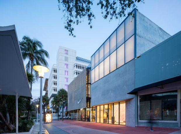 Street-level dusk view of a contemporary urban commercial building complex. The foreground facade combines large illuminated translucent window panels, exposed concrete or pale rendered walls, dark metal trim, and a projecting canopy over storefront glazing. A textured perforated or breeze-block screen and taller modern buildings are visible along a landscaped pedestrian walkway with palms and architectural street lighting.