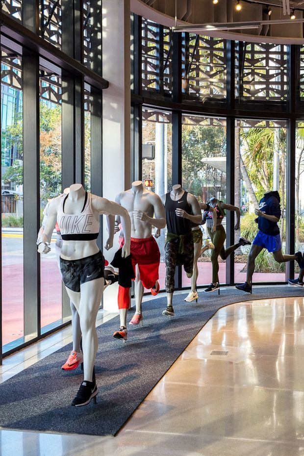 A bright, double-height retail interior with large floor-to-ceiling glazed walls and doors, displaying athletic apparel on running mannequins. The space has polished light flooring, dark window and door frames, exposed dark ceiling structure, and prominent decorative geometric screens above the glazing. Tropical landscaping and an exterior walkway are visible outside.