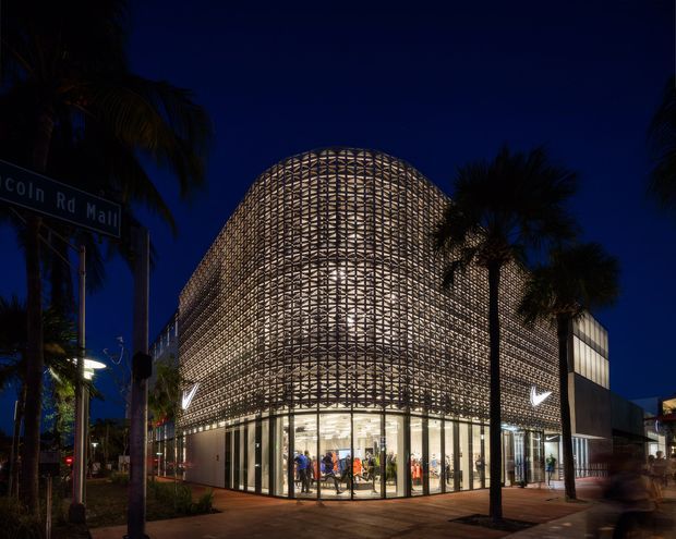 Night-time exterior photograph of a contemporary commercial building on a street corner. The illuminated ground-floor shopfront is enclosed by full-height glazing, while a dramatic curved woven screen wraps the upper facade and corner, creating a lattice-like architectural envelope. Palm trees, street signage, paving, and nighttime urban landscaping frame the building.