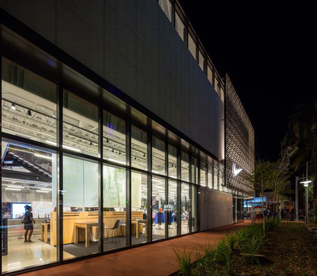 Night-time exterior view of a contemporary multi-storey commercial building. A fully glazed ground-floor frontage reveals a brightly lit retail or showroom interior with display counters and exposed services, while the upper facade is clad in dark panels and screened with a patterned perforated feature wall. A landscaped pedestrian forecourt and illuminated walkway run alongside the building.