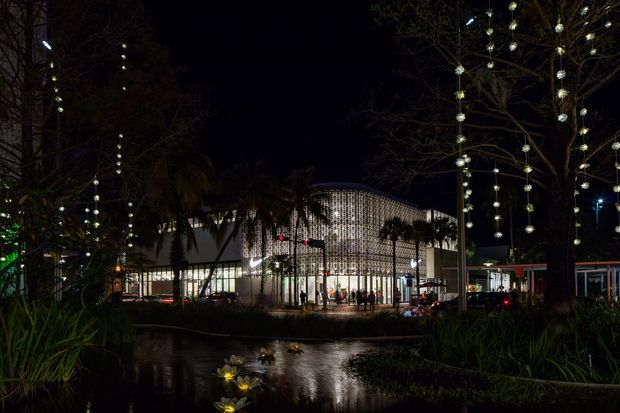 Night-time exterior view of a large contemporary pavilion or commercial building beside a landscaped pond. The building has a curved roofline and a striking perforated or patterned facade illuminated from within, with palm trees, decorative string lighting, planted garden beds, and reflections across the water in the foreground.