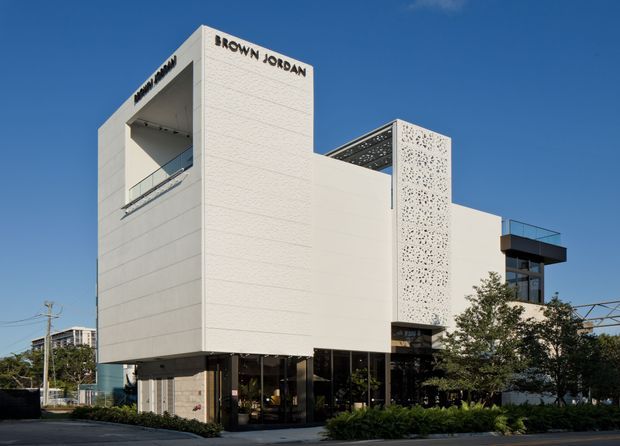 Street-facing photograph of a contemporary multi-storey commercial building with a crisp white rendered facade, deep recessed balcony, projecting box forms, black-framed ground-floor glazing, and a perforated decorative screen. The building signage reads “BROWN JORDAN,” with landscaped planting along the frontage and a clear blue sky.