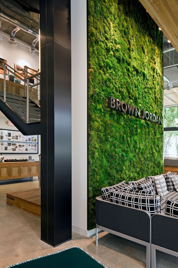 A commercial-style interior reception or lounge area with a dramatic preserved moss living wall bearing the Brown Jordan sign. Black upholstered lounge chairs with geometric patterned cushions sit on a polished concrete floor. To the left, an open industrial mezzanine with exposed services, metal stairs, and timber joinery is visible. The space combines greenery, dark metal framing, timber accents, and contemporary furnishings.
