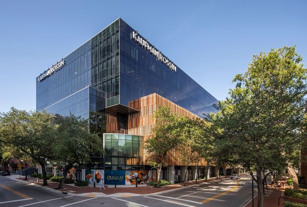 Exterior view of a contemporary multi-storey commercial building on a tree-lined urban street. The building has a large reflective blue-grey glazed facade, a projecting glazed corner volume, and a contrasting section of vertical warm timber-toned screening. Building signage is visible along the roofline and street-level frontage.