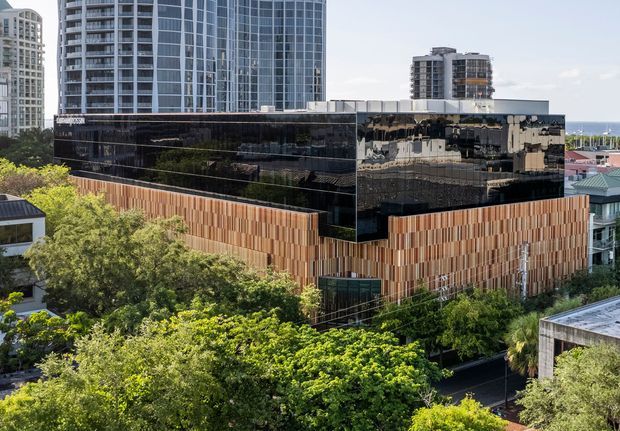 Exterior architectural photograph of a contemporary multi-storey commercial building in an urban setting. The building features a dark reflective glazed upper volume, a strongly articulated lower facade clad in narrow vertical timber-toned panels, and a flat roof. High-rise buildings and glimpses of the waterfront are visible in the background, with mature trees surrounding the site.