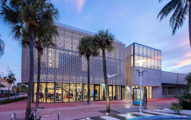 Dusk exterior view of a contemporary commercial building with a large perforated geometric facade screen, extensive glazed storefronts, a glazed upper-level corner volume, palm trees, paved plaza, and a shallow water feature in the foreground. The building appears to be a retail or mixed-use commercial space.