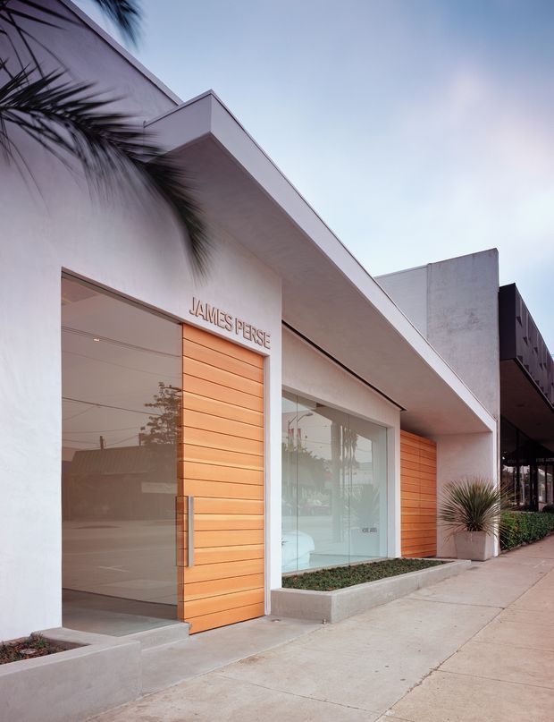 Street-facing contemporary commercial facade for a James Perse retail store, with a minimalist white rendered exterior, broad projecting flat roof canopy, full-height glazed shopfront, and warm horizontal timber-clad entry doors and wall panels. Low concrete planter beds and sculptural potted plants line the concrete footpath. The image is photographed from the sidewalk looking obliquely along the building frontage.