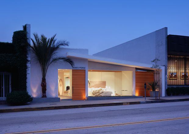 Twilight street-facing view of a sleek, low-rise modern retail or commercial facade. The building has smooth white geometric walls, a projecting flat canopy, warm-lit full-height display glazing, and a timber-toned entrance panel bearing the James Perse name. Palm trees and clipped landscaping frame the frontage, while an adjacent darker storefront is visible at right.