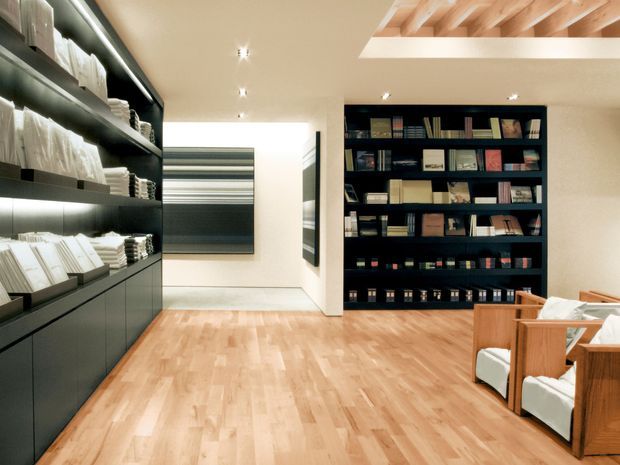 A refined library or reading room with full-height dark built-in shelving filled with books and neatly arranged objects. The room features warm timber flooring, cream walls, recessed ceiling lighting, a skylight or ceiling opening with exposed light timber beams, and timber-framed lounge chairs with white upholstery. The palette is warm and balanced, combining dark joinery with light wood and neutral finishes.