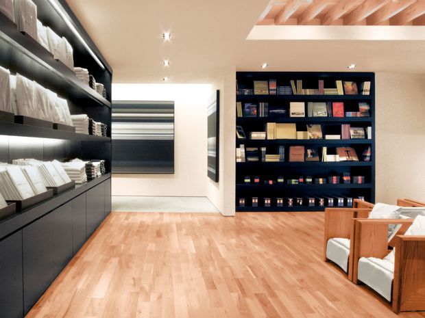 A refined, contemporary library or reading room with extensive dark built-in shelving filled with books and objects along two walls. Warm timber flooring and timber lounge chairs with light cushions contrast with the dark joinery. A large abstract artwork is visible at the rear, while a timber-lined ceiling recess adds architectural interest. Integrated shelf lighting creates a calm, curated atmosphere.