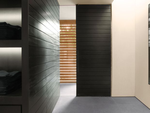 A minimalist interior passage or wardrobe area with dark horizontal timber wall panelling, a tall matching slatted door or screen, open shelving with folded textiles, warm timber venetian blinds, and a smooth light-grey floor. The space is sparsely furnished and strongly defined by built-in joinery and linear surfaces.