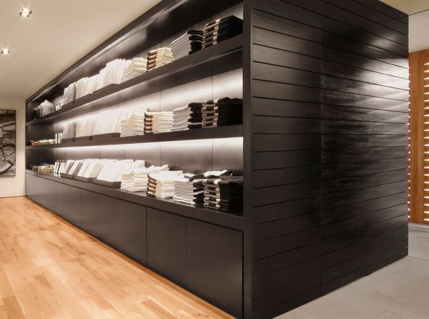A refined retail-style display area with extensive dark built-in shelving and cabinetry, neatly arranged with folded textiles and books. Warm integrated shelf lighting contrasts with the dark joinery, while light timber flooring and a dark horizontal timber feature wall create a contemporary, high-end atmosphere. A partial adjacent space with vertical timber screening is visible at right.