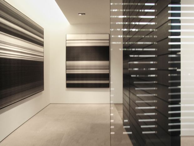 Minimalist contemporary interior corridor or gallery-like space with warm off-white walls, a pale smooth tiled or stone floor, and large abstract monochrome artworks. A full-height dark reflective glazed or glossy partition on the right creates strong horizontal reflections and visual depth. Recessed ceiling lighting and clean flush detailing reinforce the refined, restrained design.