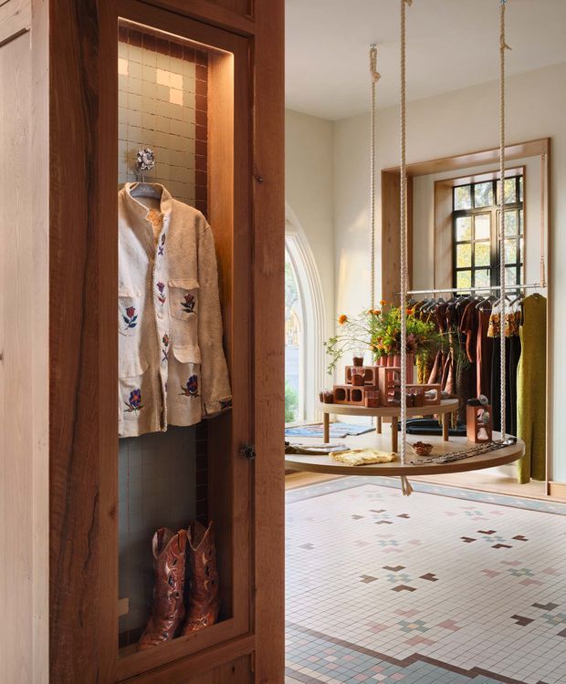 A warm, richly detailed dressing and wardrobe area viewed through a tall dark timber display cabinet. The cabinet has a glazed or open niche lined with small square tiles, displaying a shirt and boots. Beyond it is a bright retail-like dressing space with a suspended circular timber clothing rack, hanging garments, display shelving, a decorative leaded-glass window, and a patterned small-format tiled floor. The space combines custom timber joinery, warm lighting, and vintage-inspired detailing.