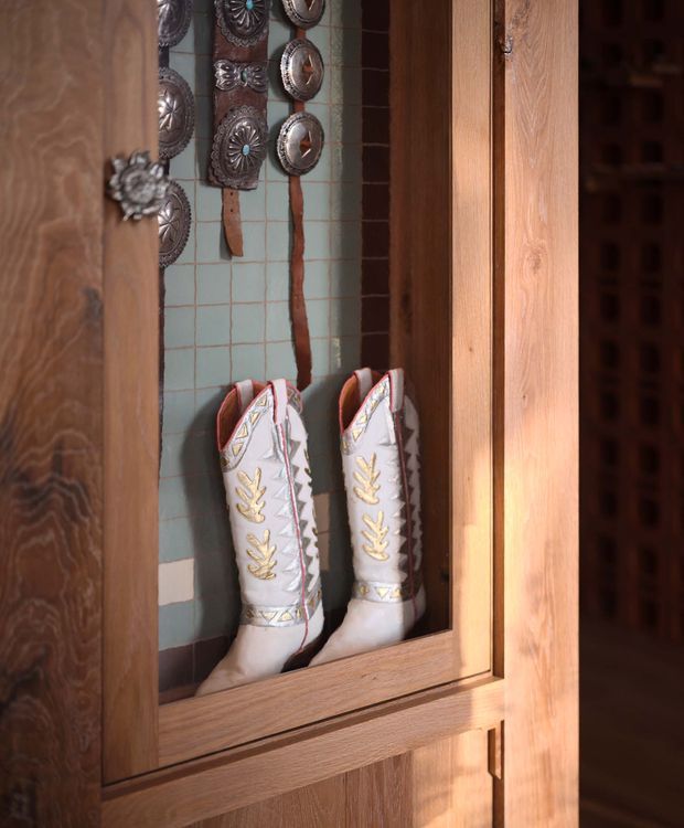 A close-up view of a tall timber-framed display cabinet or wardrobe with glass doors. Inside, a pair of decorative white embroidered boots stands against a muted turquoise tiled backing, with ornate metal medallions and leather straps displayed above. The cabinet has warm natural wood framing and detailed traditional decorative contents.