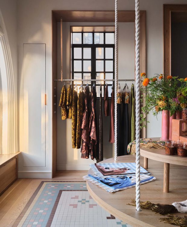 A warmly lit, fashion-oriented dressing and display space with hanging garments arranged on a rail in front of a dark framed grid window. Built-in or freestanding timber-toned joinery and a curved wooden table occupy the foreground, with magazines and decorative objects on top. Thick white rope elements hang vertically through the room, while a colourful geometric rug adds pattern over the timber floor. The space blends refined contemporary interiors with eclectic, artisanal styling.