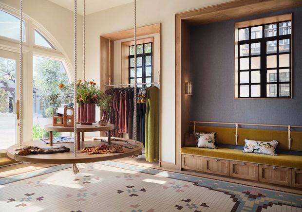 Bright, carefully styled retail or showroom interior with large arched glazing, black-framed windows, warm timber display niches, clothing racks, a central circular display table, built-in cabinetry and an upholstered window seat. The floor features a patterned geometric tile finish, while the palette combines cream walls, natural timber, charcoal grey, mustard upholstery and muted accents.