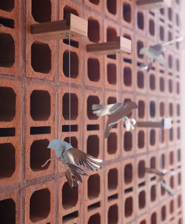 Close-up architectural detail of a perforated terracotta brick screen wall with deep rectangular openings. Small timber boxes project from the wall and suspend sculptural metal bird forms on chains, creating a playful decorative installation. The image is tightly focused on the fixed masonry facade and its texture, with shallow depth of field toward the background.