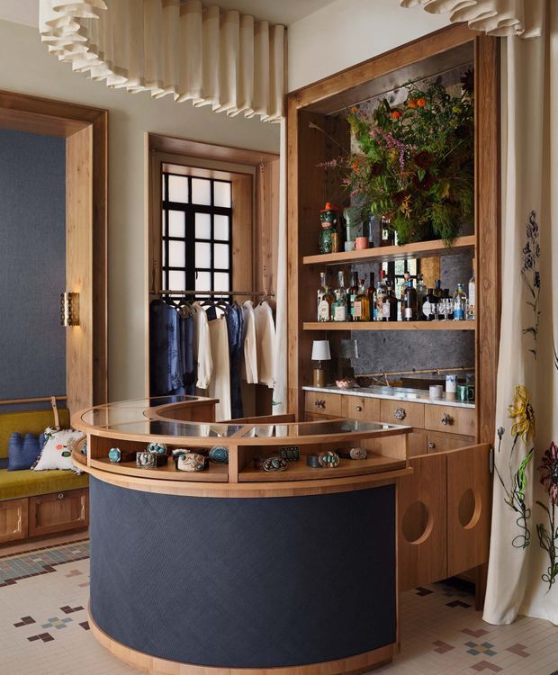 A richly detailed boutique-style retail interior featuring a curved central display counter with dark blue panelled sides and timber edging, showcasing jewellery or small accessories beneath a glass top. Behind it is a timber-framed built-in shelving and cabinetry unit with bottles, objects, and a large floral display. The room includes warm timber trim, patterned mosaic tile flooring, deep blue wall panels, decorative curtains, and a partially visible clothing display. Sculptural layered ceiling elements and period-inspired window detailing add character.