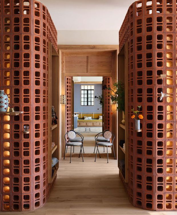 A warm, highly distinctive interior passage framed by curved screens of exposed terracotta breeze blocks with repeating rectangular openings. Pale timber flooring and a timber-lined doorway lead through to a bright living area with a black-framed window, built-in yellow seating, plants, and two small upholstered chairs in the foreground. The perforated brickwork creates strong texture, filtered light, and visual separation between spaces.