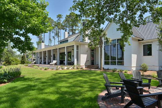 A bright exterior view of a contemporary single-storey house set within a landscaped garden. The home features pale rendered and vertical-clad walls, dark standing-seam metal gabled roofs, large black-framed windows, and a long covered veranda. A brick-paved outdoor seating area with Adirondack-style chairs occupies the foreground, surrounded by mature trees and lawn.