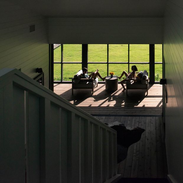 A dimly lit stairway descends toward a bright timber-framed outdoor deck. Two people sit in lounge chairs around a small round table beside expansive black-framed glazing, with a grassy landscape visible beyond. The interior stair walls and ceiling are clad in horizontal painted timber boards, while the deck uses timber flooring and opens to the outdoors.