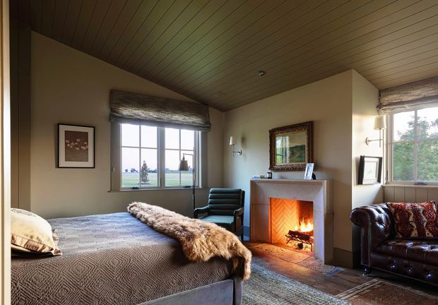 A warm, rustic bedroom with a large bed, wood-lined sloped ceiling, pale plaster walls, traditional multi-pane windows, a stone or plaster fireplace with a lit fire, leather seating, patterned textiles, and framed artwork. The room has a cozy, country-inspired atmosphere and views through the windows to an open landscape.