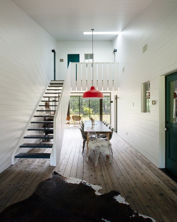 A bright, narrow double-height dining and circulation space with white horizontal wall and ceiling panelling, a timber plank floor, and an open-riser black metal staircase along the left wall. A long dining table with mixed chairs sits beneath a large red pendant light, while a white slatted balustrade overlooks the room from above. Dark green doors and trim add contrast, and a large cowhide rug occupies the foreground.