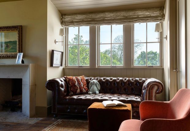 A warm, traditional sitting room with a deep brown tufted leather Chesterfield sofa positioned in front of a large multi-pane bay window. The room features a pale neutral fireplace surround, dark timber flooring, wall-mounted lamps, framed artwork, a soft upholstered pink armchair, and a brown ottoman. A raised ceiling with light timber panelling and a fabric Roman blind add architectural and textural detail.