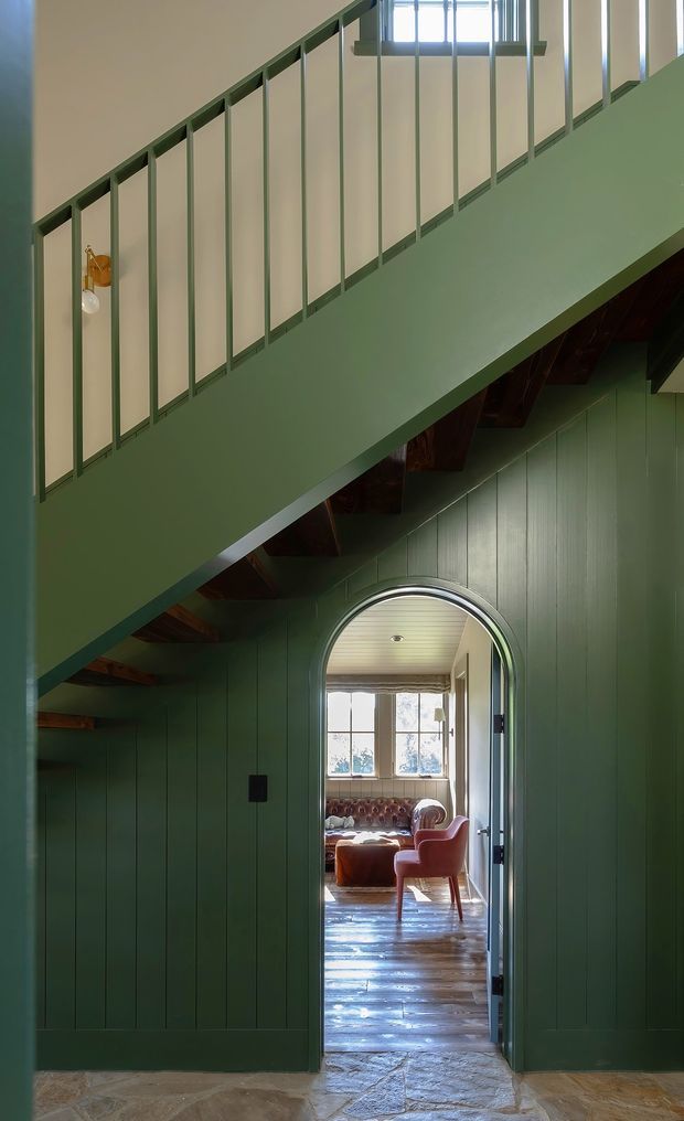 Interior architectural photograph looking beneath a staircase toward an arched doorway and a sunlit living room beyond. The stair underside and surrounding walls are clad in deep green vertical timber panelling, with a slim dark green metal balustrade above. Exposed dark timber stair structure, a small wall sconce, stone-like entry flooring, and polished timber flooring in the adjoining room are visible. The composition emphasises the stairway, arched opening, and layered circulation through the home.