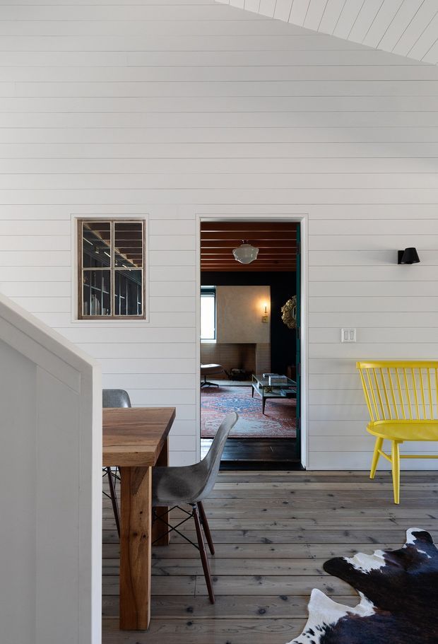 A bright, high-ceilinged interior landing or open-plan circulation space with white horizontal wall panelling and a matching angled stair balustrade. A timber dining table and dark molded chairs sit in the foreground on a wide-plank timber floor, alongside a vivid yellow spindle-back chair and a black-and-white cowhide rug. A doorway opens to a darker, richly furnished living room with a patterned rug, dark timber floor, leather seating and a wood-lined ceiling. The space combines crisp white architectural surfaces with warm timber and bold accent furniture.
