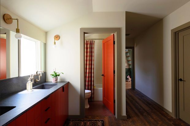 A warm, eclectic bathroom vanity area opening onto a hallway. The room features a long dark stone benchtop, reddish wood cabinetry, a large mirror, wall-mounted brass-toned lights, and a window providing natural light. An open bright orange-red door reveals a second bathroom area with a toilet and patterned shower curtain. Dark timber flooring and a patterned runner continue through the hall.