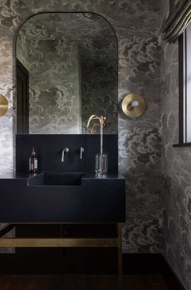 A moody, luxurious powder room with dark patterned wallcovering, a large arched mirror, matte black floating vanity and integrated basin, wall-mounted chrome tapware, and warm brass wall sconces. Dark timber flooring and black cabinetry reinforce the dramatic, refined atmosphere.