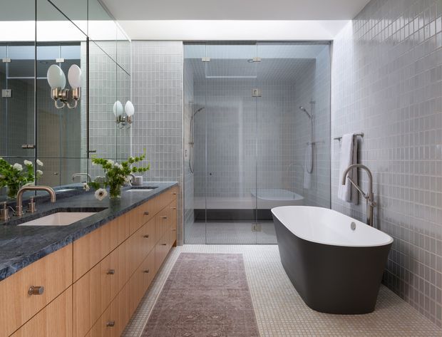 Contemporary bathroom with a double vanity featuring warm timber drawers and a dark stone benchtop, large mirrored wall, twin globe sconces, a glass-enclosed tiled shower, and a freestanding sculptural bathtub. Small-format pale grey wall and floor tiles create a clean, refined finish, complemented by a muted runner and greenery.