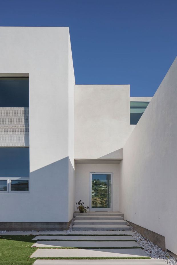 Exterior view of a minimalist contemporary residence with crisp white stucco volumes, flat rooflines, deep geometric recesses, dark-framed glazing, and a sheltered entry reached by broad concrete stepping slabs through a narrow landscaped approach. Strong sunlight creates angular shadows across the facade, with gravel edging and clipped lawn visible in the foreground.