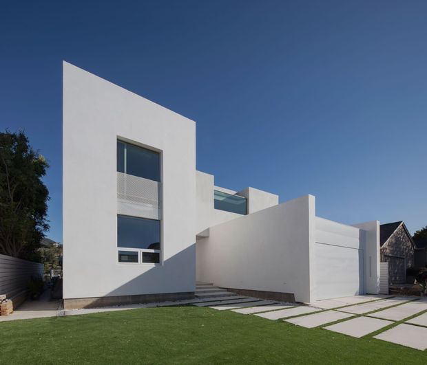 A minimalist modern multi-level house viewed from the street, with crisp white rendered geometric walls, asymmetrical rooflines, large dark-framed windows, and an integrated white garage. Pale rectangular concrete paving slabs are separated by strips of lawn, while a darker timber-clad neighbouring structure and mature planting sit at the edges of the image.