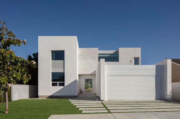 Front exterior view of a contemporary two-storey residence with a crisp white rendered facade, strong rectilinear volumes, narrow horizontal glazing, a prominent upper-level window, and an integrated double garage. Stepping-stone paving crosses the lawn toward the recessed entry, with horizontal white fencing and mature planting at the sides under a clear blue sky.
