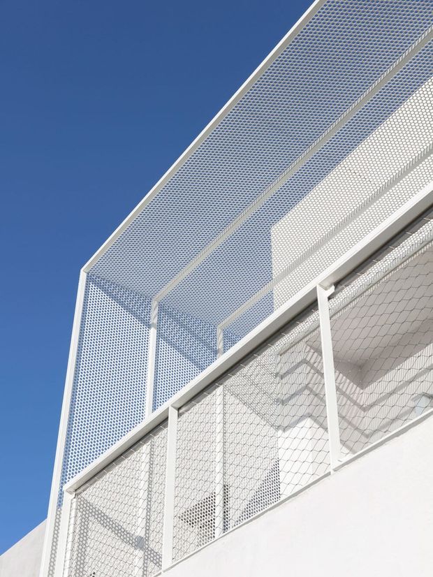 Upward-looking close-up of a contemporary white building facade featuring extensive perforated metal screening and mesh railings. The geometric screens create layered shadows against a vivid blue sky, with white structural framing and a crisp, minimalist appearance.