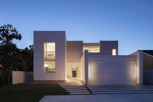 Twilight exterior view of a contemporary multi-level residence with a minimalist, rectilinear composition. The facade combines smooth dark grey and pale rendered volumes, tall narrow windows, a recessed illuminated entry, and an integrated double garage. Horizontal fencing, lawn, and broad linear concrete pavers form the foreground approach.