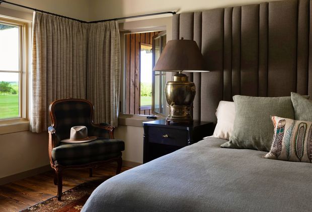A warmly lit traditional bedroom with a large upholstered bed, tall dark padded headboard, layered neutral bedding, patterned accent cushions, and a classic leather armchair with a hat. Floor-length textured curtains frame the windows, while a dark bedside chest and brass-toned table lamp add an elegant, refined character. Rural greenery is visible through the windows.