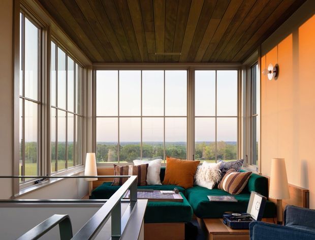 A warm, light-filled sunroom or enclosed lounge with extensive floor-to-ceiling gridded glazing overlooking a green landscape. The room features a rich timber-lined ceiling and window frames, a deep teal sectional sofa with orange, cream, and patterned cushions, soft table lamps, and a dark metal-and-glass balustrade in the foreground. The space has a relaxed mid-century-inspired character with strong indoor-outdoor connection.