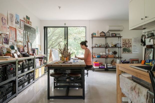 Bright, eclectic creative studio or home art workspace with a central worktable, stools, extensive open shelving, craft supplies, artwork, books, and storage baskets. Large black-framed window provides leafy views, while cabinetry and a coffee-making area occupy the right side. The room has polished light flooring, white walls, and an informal, highly personalised workspace atmosphere.