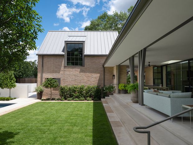 Exterior view of a contemporary brick-and-metal-roof residence arranged around a manicured courtyard lawn. The image shows a gabled brick wing with a large dark-framed window and a low, projecting glazed living area with a covered terrace, outdoor seating, steps, and a handrail. Landscaping and a pale paved pool or courtyard edge are visible.