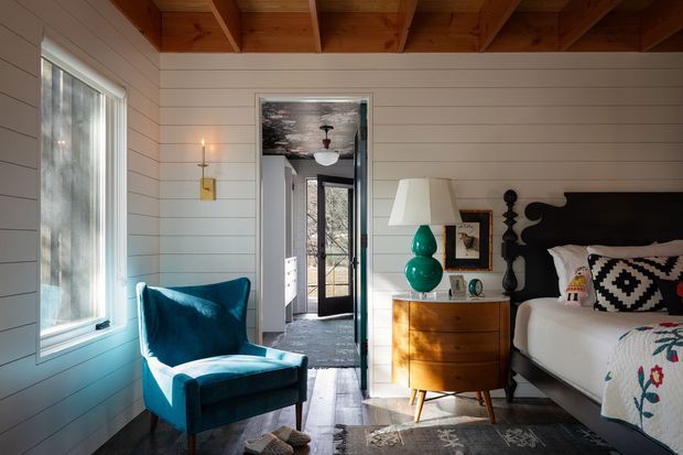 Warm, characterful bedroom with white horizontal wall panelling, exposed timber ceiling beams, dark wood flooring, and a sculptural black headboard. A teal velvet armchair sits beside a large window, while a mid-century timber dresser with a turquoise lamp adds colour. The open doorway reveals a darker adjoining circulation space and exterior glazing.