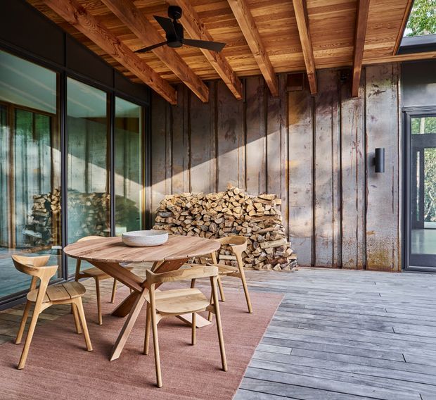 A warm, rustic-modern dining area beneath a covered timber veranda or sunroom-like enclosure. The space features a round solid-wood dining table with matching timber chairs, a stacked firewood feature against a dark vertical timber wall, exposed timber beams and rafters, large dark-framed glazed doors, grey timber flooring, and a ceiling fan. The setting has a strong indoor-outdoor connection and a natural, cabin-inspired character.