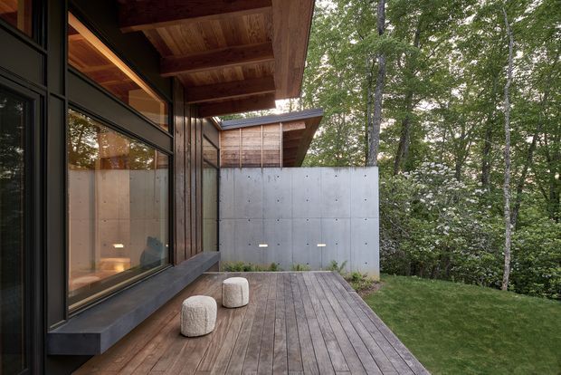 A modern timber deck and covered outdoor terrace beside a building in a wooded setting. The scene features dark-framed floor-to-ceiling glazing, warm timber soffits and decking, a projecting roof, a smooth concrete privacy wall, and two small rounded stools. Dense trees and a lawn surround the terrace.