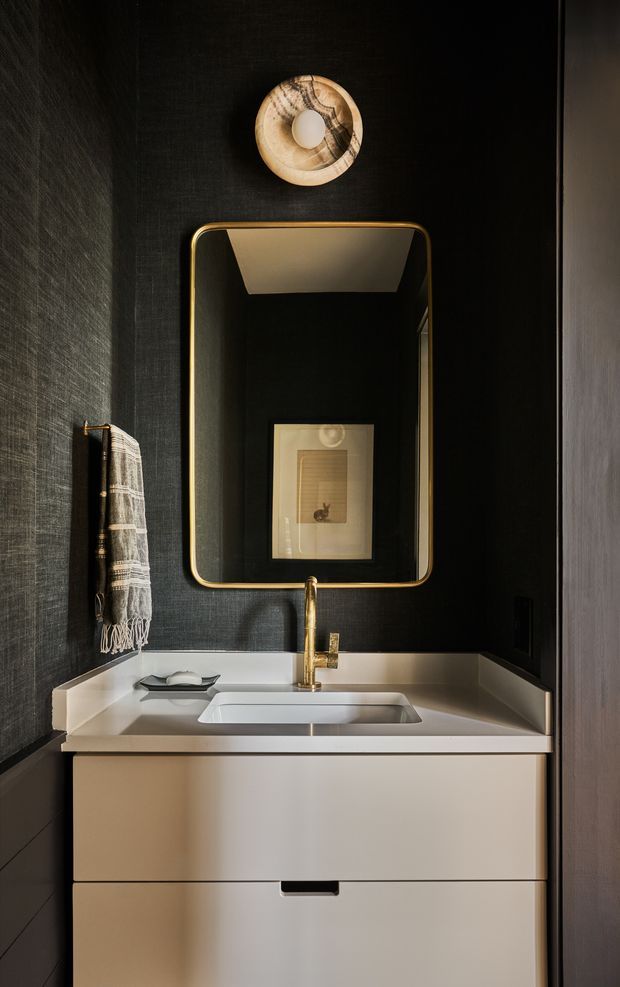 A compact, dramatically styled bathroom vanity with a white integrated basin and streamlined white cabinetry. The walls are clad in dark textured finish, contrasted by a tall rounded-rectangle mirror with a brass-gold frame and matching brass tapware. A small wall-mounted light and hanging textile add detail, while the mirror reflects framed artwork.